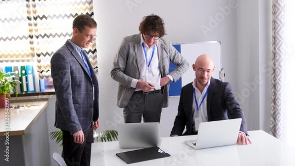 Fototapeta Three men are standing around a table with laptops and discussing something. The scene is professional and focused