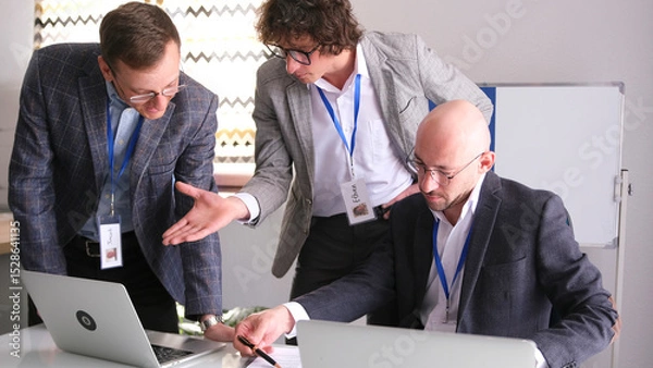 Fototapeta Three men in suits and glasses are conferring in a conference room. Businessmen are looking at laptops and working on a common project