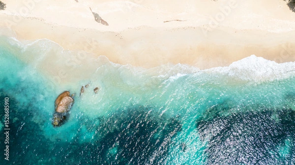 Fototapeta Aerial view of gentle turquoise waves brushing the sandy shore with a glimpse of rocks beneath the water. Praslin, Seychelles.