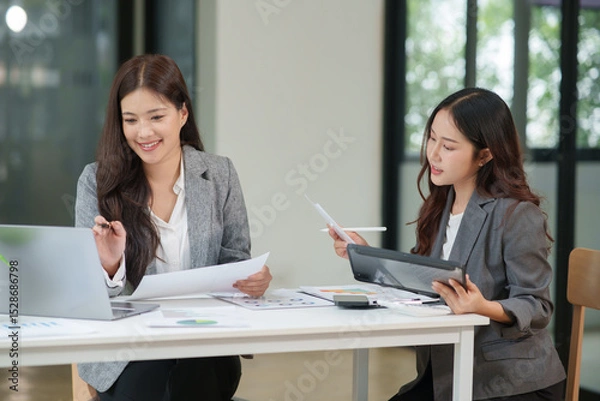 Fototapeta Two Asian businesswomen are sitting in a meeting, planning investment plans, business strategies. On the table are documents and graphs.