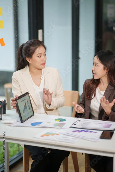 Fototapeta Two Asian businesswomen are sitting in a meeting, planning investment plans, business strategies. On the table are documents and graphs.