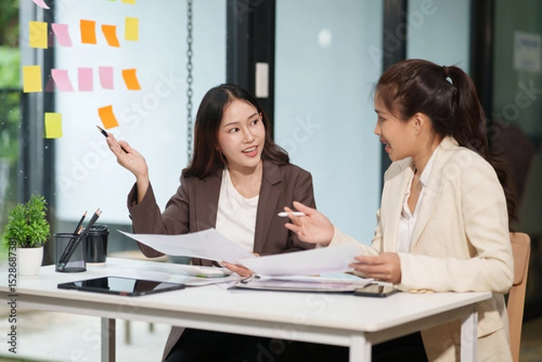 Fototapeta Two Asian businesswomen are sitting in a meeting, planning investment plans, business strategies. On the table are documents and graphs.