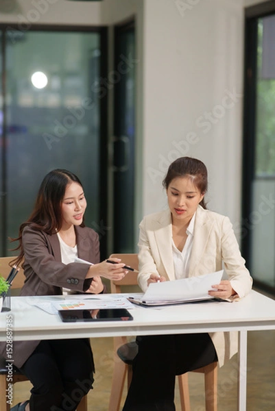 Fototapeta Two Asian businesswomen are sitting in a meeting, planning investment plans, business strategies. On the table are documents and graphs.