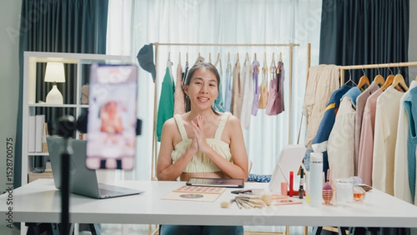 Fototapeta Young beautiful Asia lady blogger look at phone camera recording swatch apply lipstick on arm show follower review product vlog video live streaming at home. Business online social media concept.