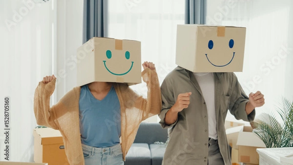 Fototapeta Happy Asia couple stand front of sofa couch used carton box made smiling face funny dance front of camera together in living room at new house. Husband and wife moving new house, Couple goal Concept.