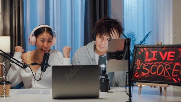 Fototapeta Young happy Asian couple wear headphone sit on desk online live streaming football live score looking at camera watch soccer sport game on laptop talk with follower in small studio at home at night.