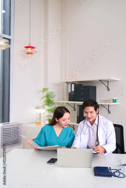 Obraz Asian Doctors Discussing Patient Care Examining Medical Records and Laptop at Hospital Office for Healthcare Professional Collaboration