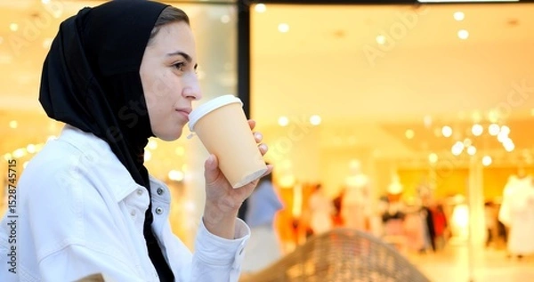 Obraz Young muslim woman wearing a hijab enjoys a coffee while using her smartphone in a brightly lit shopping mall, showcasing a blend of modern life and tradition. Free Islamic woman in a shopping center