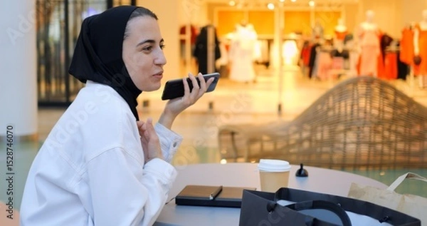 Obraz Young muslim woman wearing traditional hijab sitting at table in a shopping mall food court, talking on mobile phone and drinking coffee, enjoying a break from shopping. Free from prejudice of Muslim