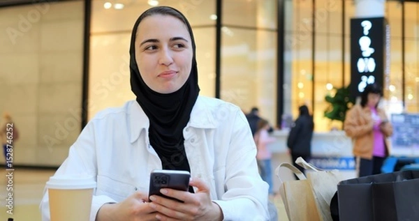 Obraz Young muslim woman wearing hijab sitting at table in shopping mall, comfortably browsing smartphone with relaxed smile, embodying modern digital lifestyle and urban connectivity