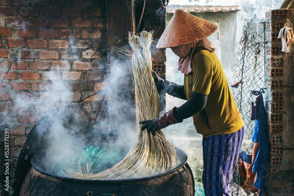 Fototapeta Vietnamese woman in a traditional straw hat dyes reeds at woven mat factory in Vietnam