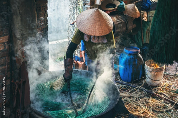 Fototapeta Vietnamese woman in a traditional straw hat dyes reeds at woven sleeping mat factory in Vietnam