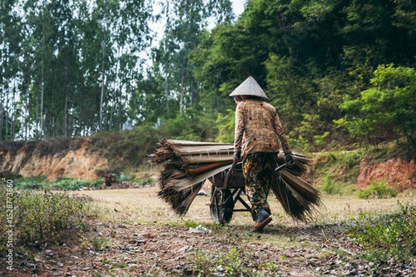 Fototapeta Vietnamese woman in traditional straw hat carries woven mats made of dyed reeds on cart at rug factory in village in Vietnam