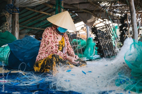Fototapeta Vietnamese woman in traditional straw hat Non La repairs fishing net in a workshop on beach in fishing village in Vietnam