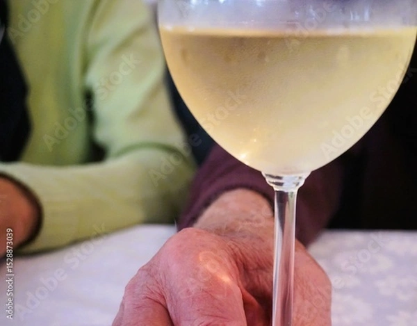 Fototapeta Hand von altem Mann umfasst Glas mit Weißwein auf Tisch mit weißer Stofftischdecke in Restaurant 