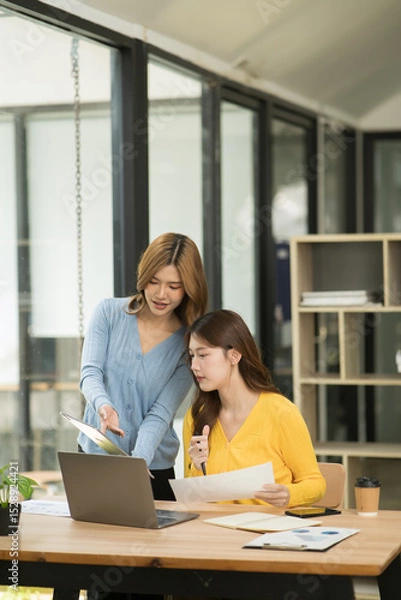 Fototapeta Two Asian businesswomen using laptops, on the table there are graph documents working on business investment.