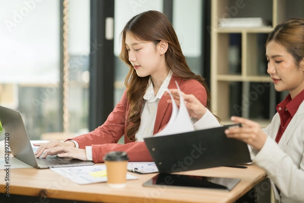 Fototapeta Two Asian businesswomen using laptops, on the table there are graph documents working on business investment.