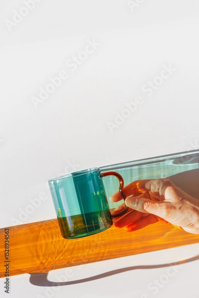 Obraz Vibrant still-life with colorful transparent glass mug or cup in shades of green teal and orange. Sunlight dynamic shadows and reflections on white, minimal aesthetic design, table setting