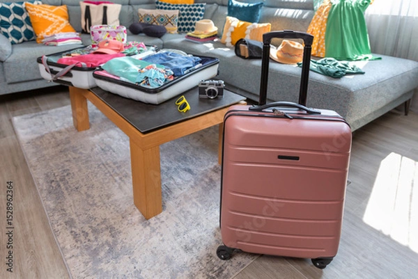 Fototapeta Luggage and clothing scattered across the sofa in a living room as part of travel preparation. Open suitcase over table with colorful summer clothes, ready for vacation. Copy space available on left.