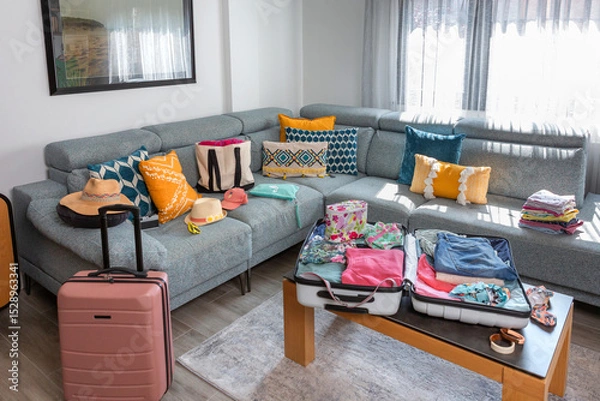 Fototapeta Open suitcase with summer clothes and accessories placed on coffee table and sofa in living room, symbolizing upcoming vacation and travel preparations