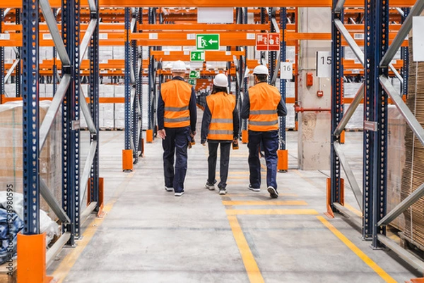 Fototapeta Warehouse workers in safety vests and hard hats navigating through a spacious logistics center, ensuring efficient operation and organization