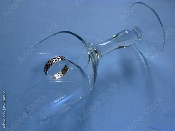 Fototapeta a broken wine glass lying on a blue surface with a silver ring hanging on a sharp glass shard. The image symbolizes relationship breakdown.
