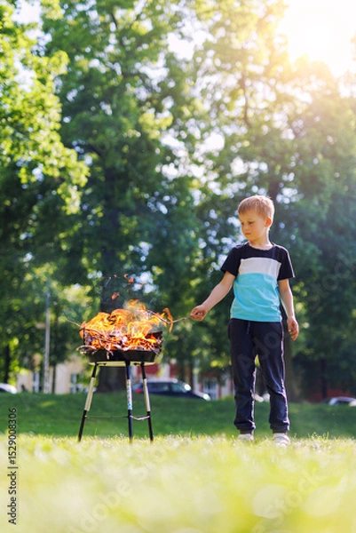 Fototapeta Cute little adorable baby boy put wooden stick in fire pit with huge fire at city park or house yard. Child play dangerous game with bonfire. Fire accident risk and health damage concept