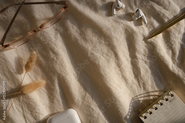 Fototapeta Glasses, wireless earbuds, pen, notebook, and dried grasses on fabric, soft light