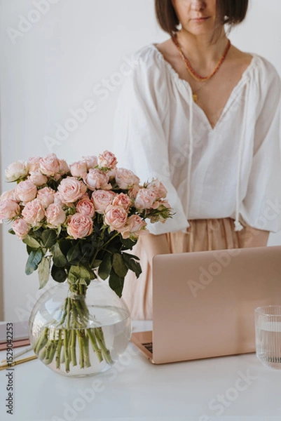 Fototapeta Elegant woman working with laptop and pink roses on desk. Aesthetic minimalist home office workspace