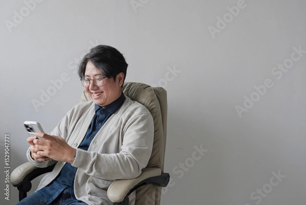 Fototapeta A man is sitting in a chair and looking at his cell phone