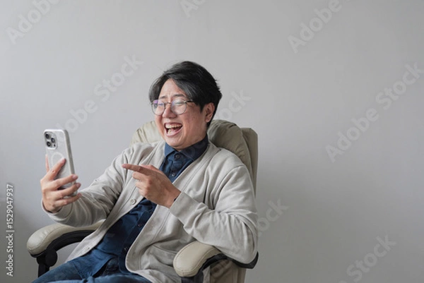 Fototapeta A man is sitting in a chair and pointing at his cell phone