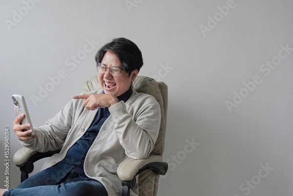 Fototapeta A man is sitting in a chair and looking at his cell phone