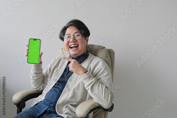 Fototapeta A man is sitting in a chair and holding a green phone