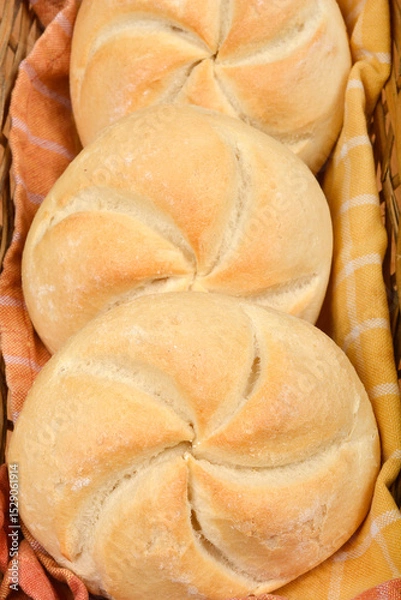 Obraz A close-up, top-down view shows three freshly baked Kaiser rolls nestled in a woven basket on a patterned cloth