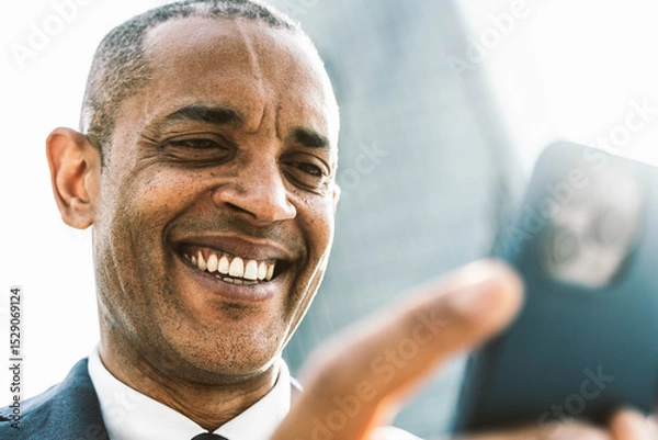 Fototapeta Smiling black businessman holding smart mobile phone in front of office building - Happy business man using online banking app on smartphone - Technology and business life style concept