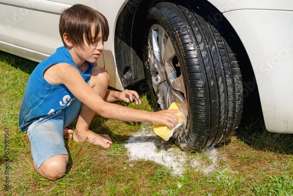 Fototapeta A boy in a blue T-shirt washes the front wheels with a sponge with foam.