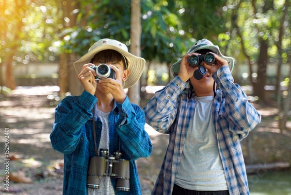 Fototapeta Two Asian boys use binoculars and a camera to study the local birds in a community forest. Soft and selective focus.