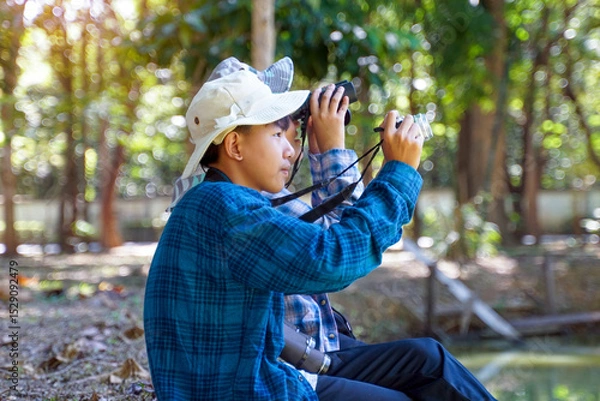 Fototapeta Two Asian boys use binoculars to study the local birds in a community forest. Soft and selective focus.