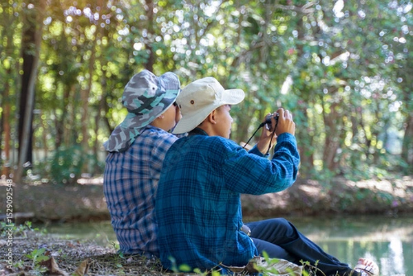 Fototapeta Two Asian boys use binoculars and a camera to study the local birds in a community forest. Soft and selective focus.