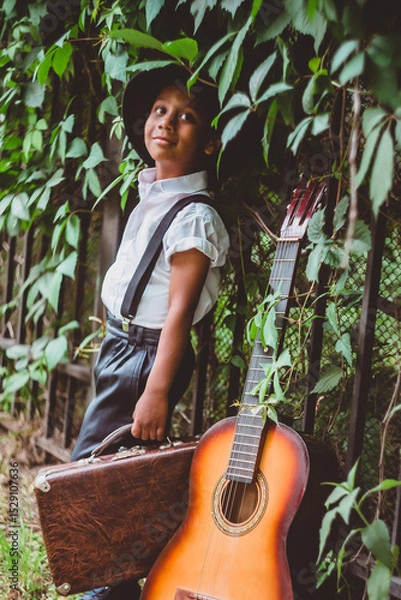 Fototapeta boy dressed in the style of the 20s stands next to a guitar holding a suitcase in his hand looking up