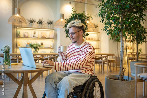 Fototapeta Interested man freelancer in wheelchair working with portable computer in cafe. Focused hipster guy student with disabilities using laptop watches webinar remote study, learning online in coffeeshop.