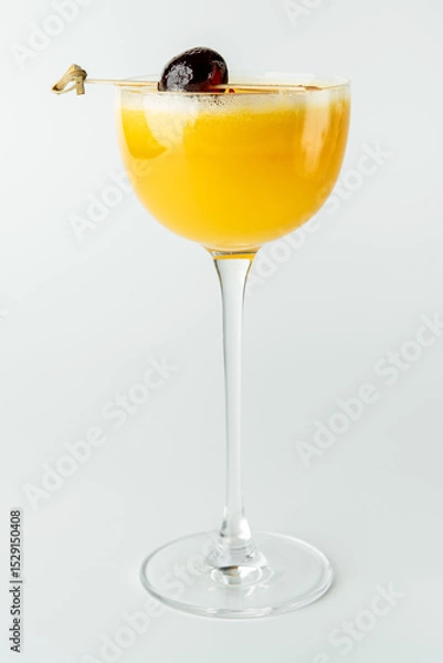 Fototapeta A vibrant yellow cocktail in a tall, elegant glass with a dark cherry garnish on a skewer, set against a clean white background, exuding a refreshing and sophisticated appeal.