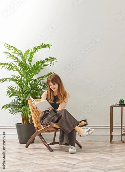 Obraz A woman is reading a book at home, looking at the phone and tablet. Decorative interior style and vase of plant.