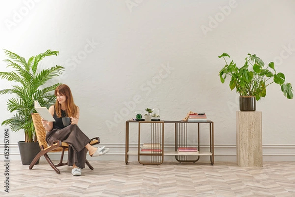 Obraz A woman is reading a book at home, looking at the phone and tablet. Decorative interior style and vase of plant.