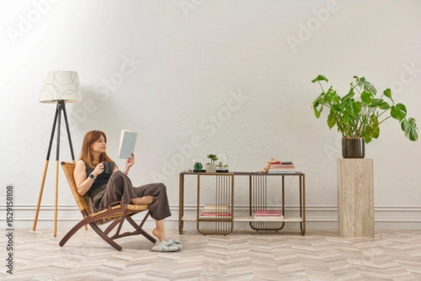 Obraz A woman is reading a book at home, looking at the phone and tablet. Decorative interior style and vase of plant.