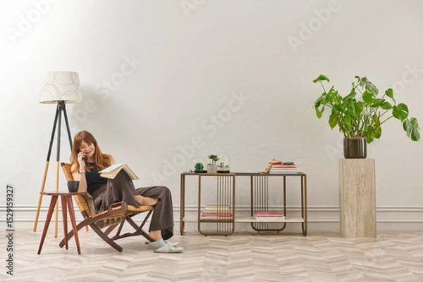 Obraz A woman is reading a book at home, looking at the phone and tablet. Decorative interior style and vase of plant.
