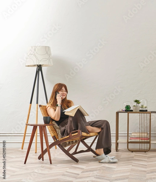 Obraz A woman is reading a book at home, looking at the phone and tablet. Decorative interior style and vase of plant.