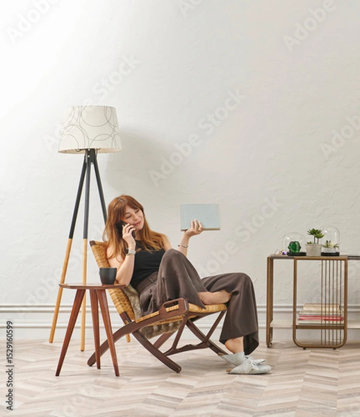 Obraz A woman is reading a book at home, looking at the phone and tablet. Decorative interior style and vase of plant.