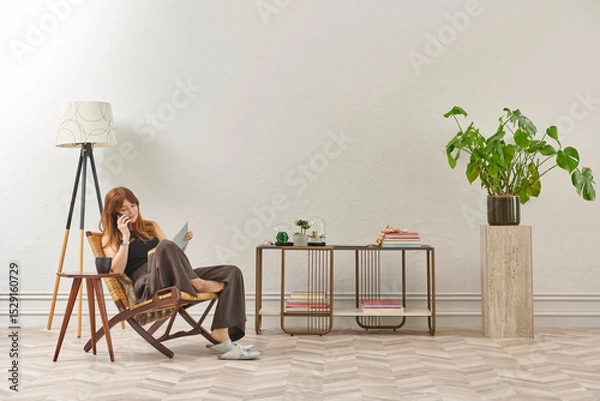 Obraz A woman is reading a book at home, looking at the phone and tablet. Decorative interior style and vase of plant.