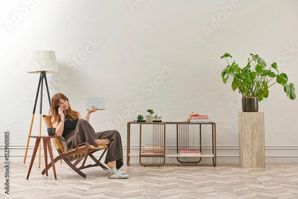 Obraz A woman is reading a book at home, looking at the phone and tablet. Decorative interior style and vase of plant.